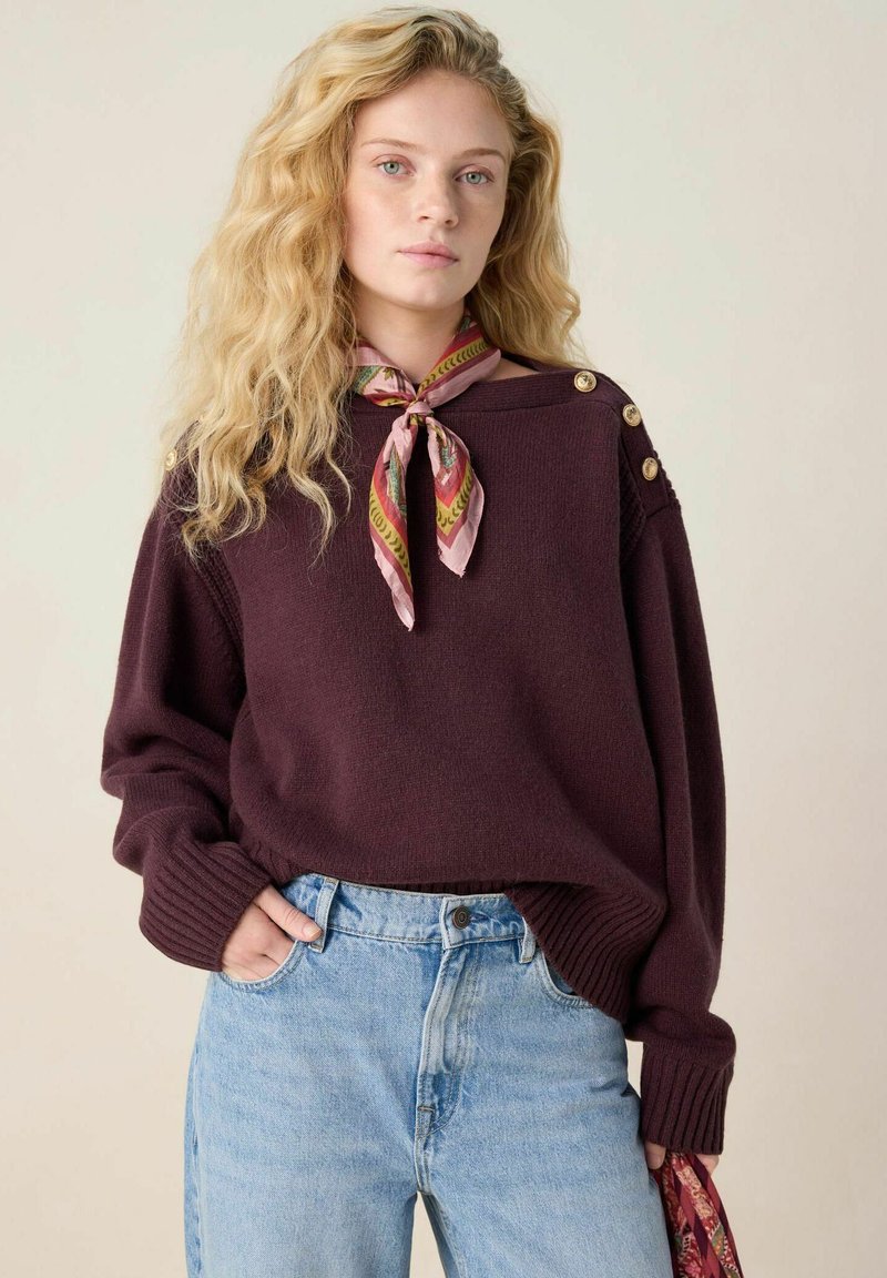 Bordeaux gebreide trui met geribbelde manchetten, voorzien van gouden knoopdetails op de schouders, gecombineerd met lichtblauwe high-waisted jeans en een roze patroon sjaal.