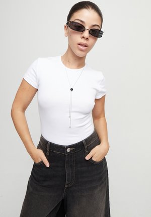 Camiseta blanca ajustada combinada con pantalones cortos de mezclilla oscuros. El modelo lleva unas pequeñas gafas de sol negras y un collar largo de plata con un colgante negro.