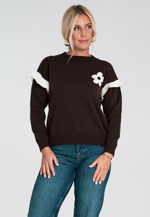 Blondynka ubrana w ciemno-brązowy sweter z białymi falbanami i białym kwiatem na piersi, w połączeniu z niebieskimi dżinsami, stoi.
