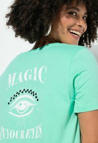 Mátově zelené tričko s krátkými rukávy, na kterém je bílý nápis "MAGIC IN YOUR EYES" a motiv oka s černými diamantovými akcenty.