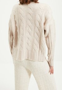 Pull en tricot torsadé de couleur beige doux, avec poignets côtelés et dos légèrement raccourci. Porté avec un pantalon texturé de couleur crème.