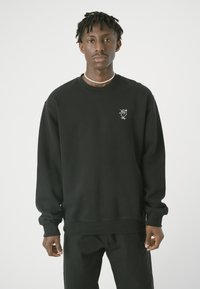 Schwarzes Sweatshirt mit Rundhalsausschnitt und langen Ärmeln. Mit einem kleinen weißen Grafikdesign auf der linken Brust. Aus Baumwollmaterial, entspannter Schnitt.