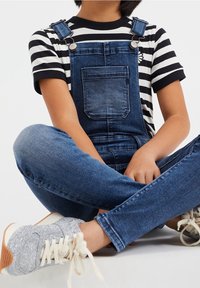 Jeansoveraller med metallspännen, med en framficka, bärs över en svart-och-vit randig t-shirt. Glittriga grå sneakers komplementerar looken.