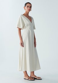 Lichtbeige jurk tot op de knie met een diepe V-hals, korte pofmouwen en een getailleerd middel. Heeft vloeiende stof en een minimalistisch ontwerp.