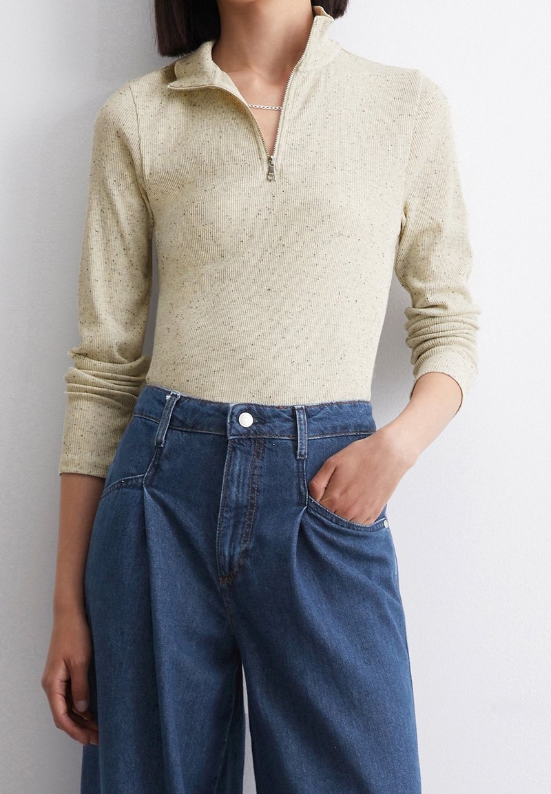 Kobieta ubrana w beżowy sweter z zamkiem błyskawicznym w cętki i niebieskie dżinsy z wysokim stanem o szerokich nogawkach, jedna ręka w kieszeni, na tle białej ściany.