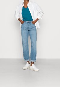 Jeans de mezclilla azul claro con pierna recta y dobladillos, combinados con una blusa de pico en teal y una camisa blanca de botones. Zapatillas blancas.
