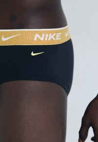 Svarta bomullsboxer kalsonger för män med en gul stickad midja som har "NIKE"-logotypen och en liten gul logotyp på framsidan.