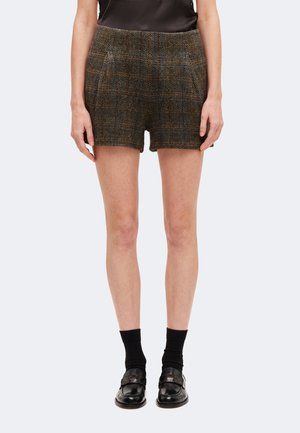 Femme portant un short à carreaux marron associé à des chaussettes noires et des mocassins en cuir noir, debout les mains détendues le long du corps.