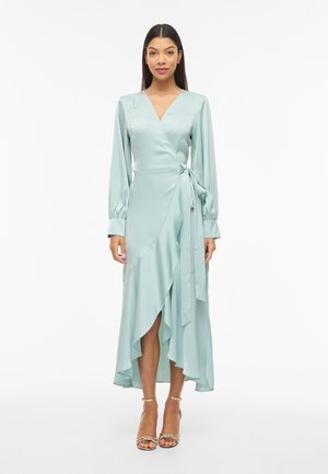 Kvinne står iført en lys blå omslagskjole med lange ermer, sidestropp og volang på asymmetrisk kant, kombinert med gullfargede ankelsnere hæler.