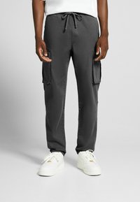 Mörkgrå cargo-byxor med dragsko i midjan, utrustade med två sidofickor och en avslappnad passform, kombinerat med vita sneakers.