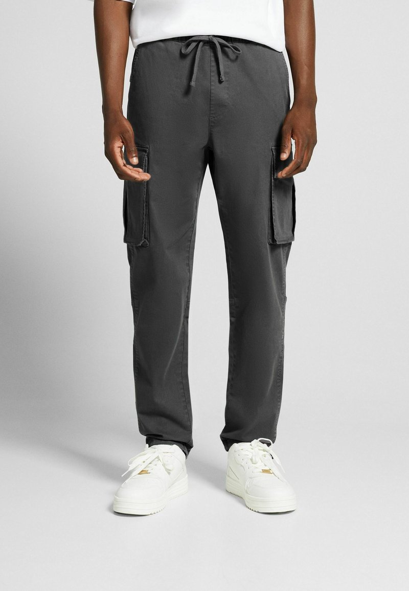 Mörkgrå cargo-byxor med dragsko i midjan, utrustade med två sidofickor och en avslappnad passform, kombinerat med vita sneakers.