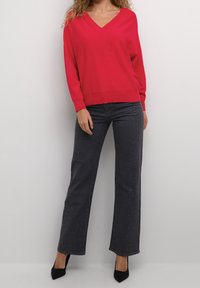 Frau in einem roten V-Ausschnitt-Pullover, dunkelgrauen, geraden Jeans mit kleinen Verzierungen und schwarzen, spitzen High Heels vor einem weißen Hintergrund.