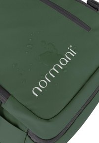 Grüne Tasche mit strukturierter Oberfläche, ausgestattet mit einer wasserabweisenden Beschichtung und einem silbernen Logo. Enthält einen schwarzen Reißverschluss und Akzente.