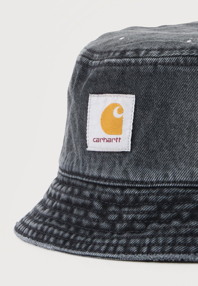 Dunkelgrauer Denim-Bucket-Hut mit einem weißen rechteckigen Stoffetikett mit gelbem Carhartt-Logo und rotem Text.