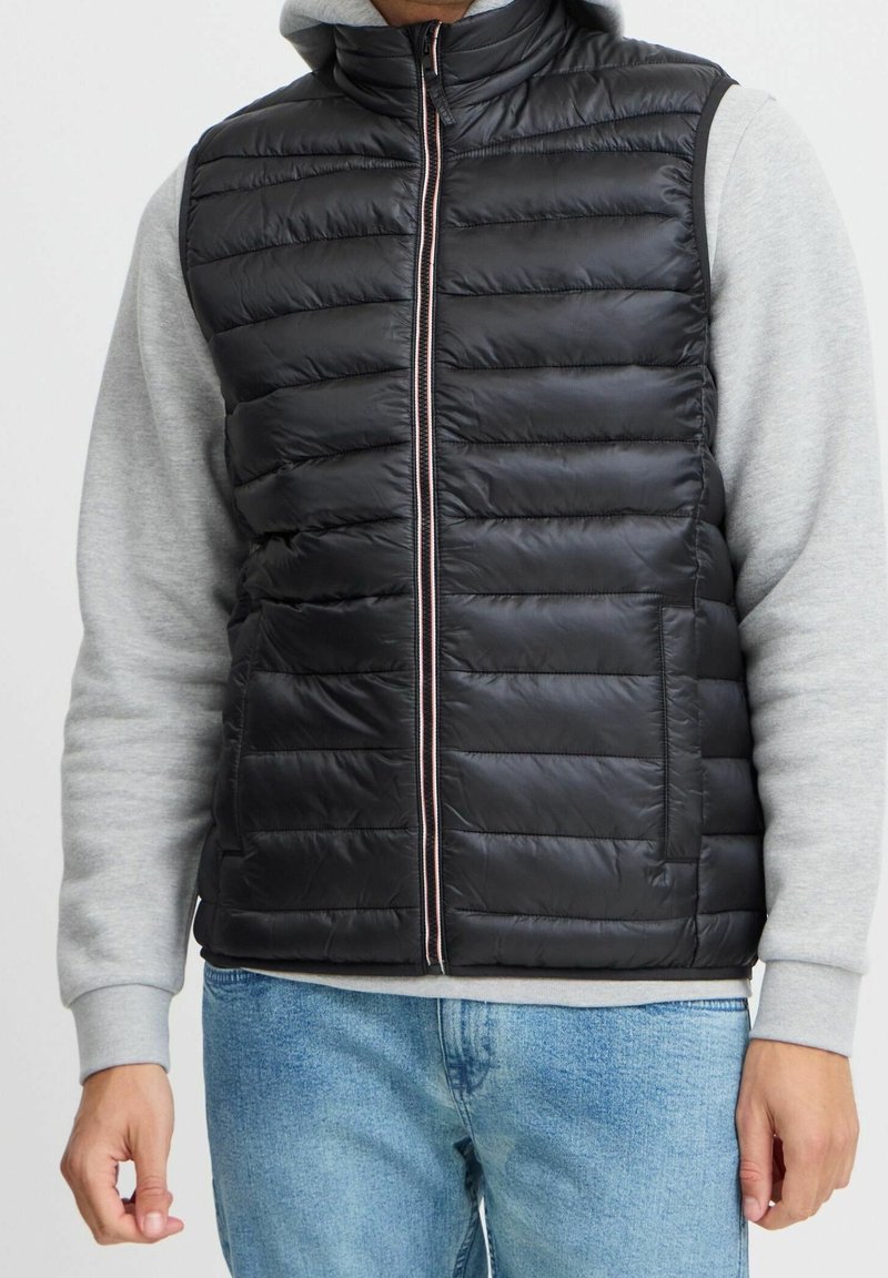 Personne portant un gilet sans manches matelassé noir par-dessus un sweat-shirt gris clair et un jean bleu clair sur un fond uni.
