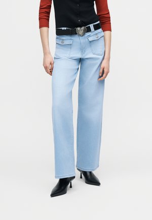 Γυναίκα που φοράει ανοιχτό γαλάζιο τζιν ψηλόμεσο με τσέπες με καπάκι μπροστά, μαύρη ζώνη με αγκράφα καρδιά, μαύρες μυτερές γόβες και κόκκινο μπλούζα με μακριά μανίκια.