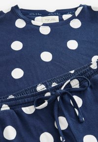 Costum format albastru marin, compus dintr-o bluză și o pereche de pantaloni, cu buline mari albe. Bluza cu mâneci scurte are un guler rotund; talia include un șnur elastic.