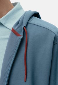 Plava kapuljača glatke teksture, s mareličastim vezicom i svijetloplavom košuljom ispod. Detalj s patentnim zatvaračem sa strane.