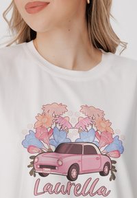 Balta medvilnė marška su rožinės spalvos automobilio grafika, apsupta spalvingų gėlių ir tekstu „Laurella“ žaismingu šriftu.