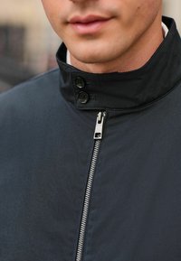 Black jacket featuring a high collar, two buttons, and a front zipper with metallic hardware. Smooth texture, minimal design.