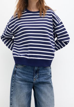 Personne portant un t-shirt à manches longues rayé horizontalement bleu marine et blanc avec un jean bleu, debout les mains derrière le dos.