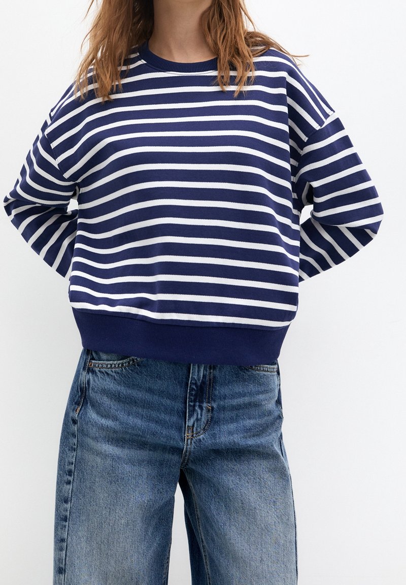 Personne portant un t-shirt à manches longues rayé horizontalement bleu marine et blanc avec un jean bleu, debout les mains derrière le dos.