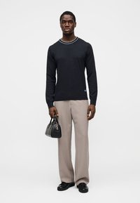 Pull en maille bleu marine avec un col rond contrastant, associé à un pantalon beige à jambes larges. Le sujet tient un petit sac à main noir.