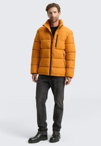 Pufferjack in feloranje met een horizontaal gestikt ontwerp, hoge kraag en een rits aan de voorkant. Gekoppeld aan donkere jeans en zwarte laarzen.
