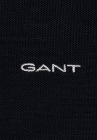 Schwarzer, strukturierter Stoff mit einem hervorgehobenen weißen, gestickten Logo, das "GANT" liest. Das Design ist einfach und konzentriert sich auf die Markenidentität.