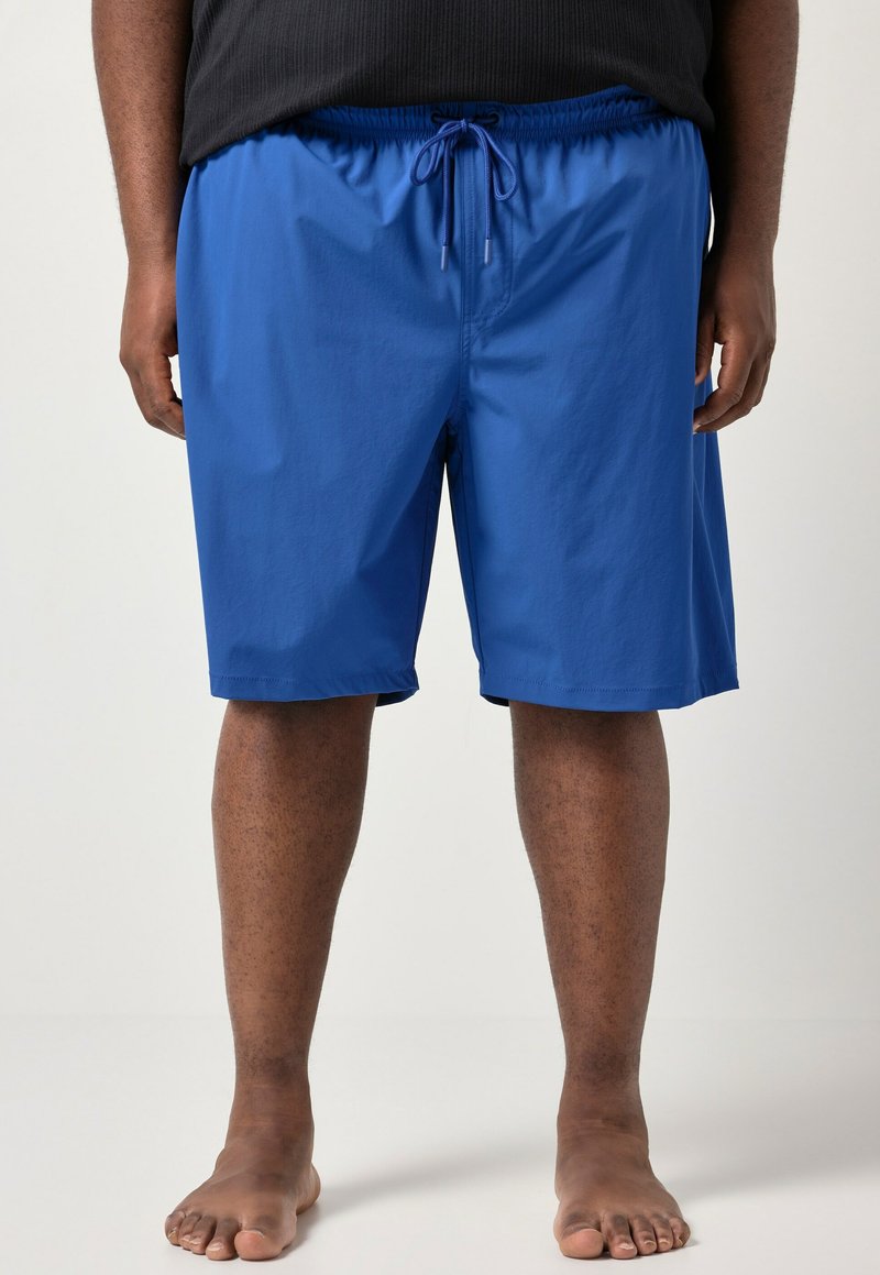 Hombre con pantalones cortos azules con cordón y camiseta negra, de pie descalzo sobre un fondo claro y liso.