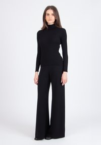 Juodo, matinio trikotažo turtlenecko megztinis, derintas su plačiomis juodomis kelnėmis, abi pagamintos iš minkštos medžiagos. Modelis vilki juodus batus.