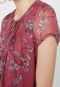 Bordová květinová blůza s krátkými, mírně volánkovými rukávy. Materiál je lehký a průhledný, s modrými květinovými vzory na pozadí.