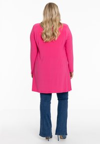 Langemouwen, felroze tuniek met een rechte snit, tot halverwege de dij, gecombineerd met blauwe flared jeans en hoge hakken. Glad textiel.