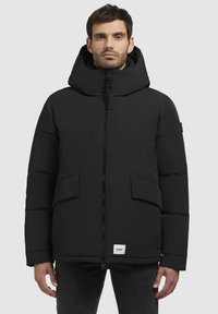 Sort pufferjakke med hætte, frontlynlås og to store sidelommer. Har et logo-tag nederst. Glat tekstur og strømlinet design.