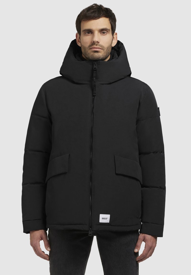 Sort pufferjakke med hætte, frontlynlås og to store sidelommer. Har et logo-tag nederst. Glat tekstur og strømlinet design.