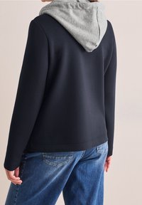 Personne portant un sweat à capuche noir avec une capuche grise et un jean bleu ample, debout dos à la caméra.