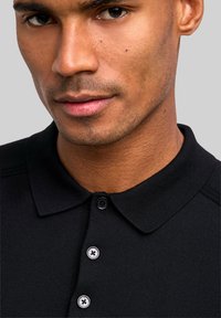 Czarna koszulka polo z kołnierzykiem, trzema czarnymi guzikami i fakturowaną tkaniną. Zbliżenie na koszulkę na szyi i twarzy modela.