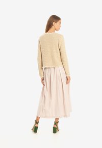 Beige, texturerad jumper med fransar, bärs över en lång rosa kjol. Gröna klacksandaler med blockklack kompletterar outfiten.