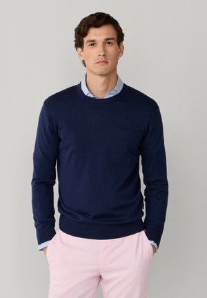 Giovane uomo che indossa un maglione blu navy sopra una camicia a righe blu con pantaloni rosa chiaro, in piedi contro uno sfondo grigio chiaro uniforme.