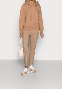 Sudadera marrón con cordones, combinada con pantalones slim-fit beiges claros. Se lleva con zapatillas blancas y beiges y un pequeño bolso texturizado de color crema.