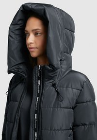 Black, puffy down jacket with a large hood, featuring a zippered front and drawstring accents. Textured fabric with visible quilted pattern.