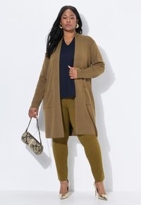 Eine Frau trägt einen langen braunen Cardigan, ein marineblaues Oberteil, olivfarbene Hose, goldene Ohrringe und High Heels, während sie eine kleine Handtasche mit Schlangenledermuster hält.