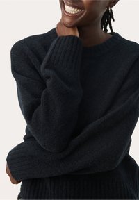 Personne souriante portant un pull en maille texturée foncée avec des poignets côtelés, tenant son cou d'une main, cadré à partir du nez vers le haut.