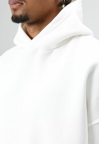 Sweat à capuche blanc avec une texture douce, doté d'un col montant, de bords bruts et d'une coupe décontractée. Aucun logo ou motif visible.