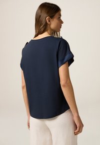Top de manga corta azul marino con cuello redondo y puños enrollados. Tejido suave con un corte relajado. Presenta un botón decorativo en el hombro.
