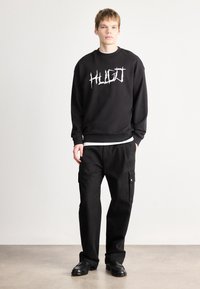Μαύρο φούτερ με λευκό λογότυπο "HUGO", συνδυασμένο με χαλαρές μαύρες cargo παντελόνες με πλευρικές τσέπες. Μοντέλο που φοράει μαύρα παπούτσια.