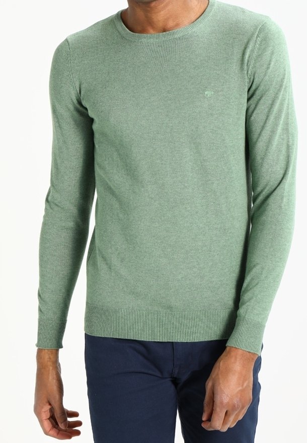 Homme portant un pull vert ajusté à manches longues avec des poignets et un ourlet côtelés, associé à un pantalon bleu foncé, debout devant un fond blanc.