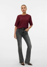 T-shirt bordeaux à manches courtes associé à un jean gris taille haute. Le jean a une jambe évasée et le modèle porte des talons ornés de couleur brune.