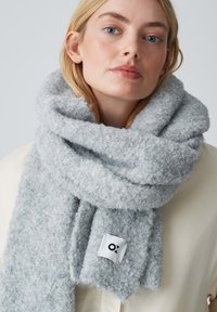 Grijze, fluffy sjaal gemaakt van zacht materiaal, met een wit label waarop de letters 'O' en 'X' staan aan één uiteinde, om de nek gedrapeerd.