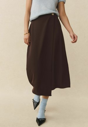 Frau trägt einen braunen Midirock mit einem goldenen Knopf, ein hellblaues ärmelloses Oberteil, hellblaue Socken und schwarze spitze Schuhe.